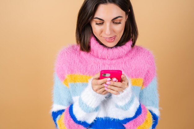 Young woman on beige in a knitted sweater with a phone looks at the screen and smiles licks her lip