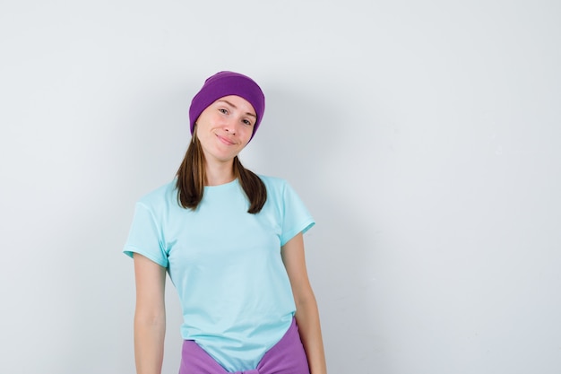 Young woman bending head while posing in blue t-shirt, purple beanie and looking cheery , front view.