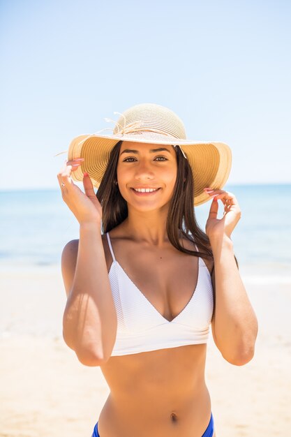 Young woman in bikini wearing white straw hat enjoying summer vacation at beach. Portrait of beautiful latin woman relaxing at beach with sunglasses.