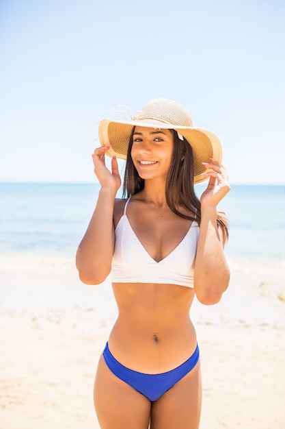 Young woman in bikini wearing white straw hat enjoying summer vacation at beach. Portrait of beautiful latin woman relaxing at beach with sunglasses.