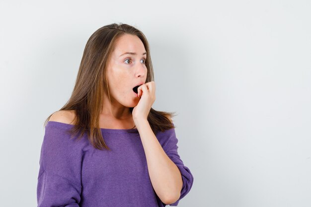 Young woman biting fist emotionally in violet shirt and looking frightened. front view.