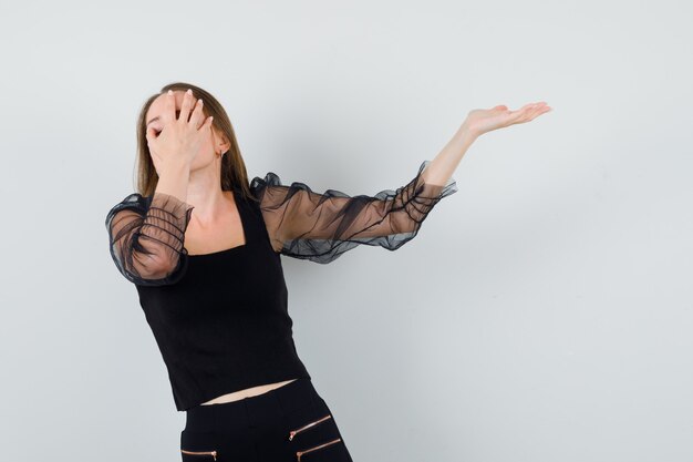 Young woman in black blouse and black pants covering face with one hand and holding another hand as presenting something and looking serious 