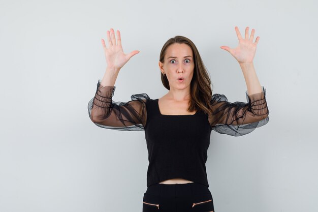 Young woman in black blouse and black pants standing in surrender pose and looking scared , front view.