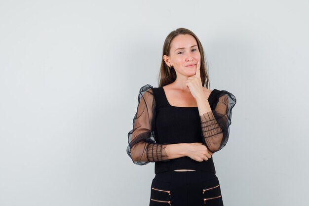 Young woman in black blouse holding hand on jaw and looking pensive 