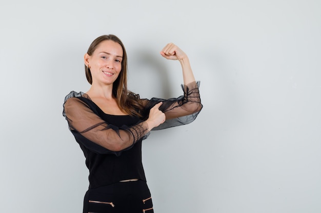 Young woman in black blouse pointing at her arm muscles and looking pleased 