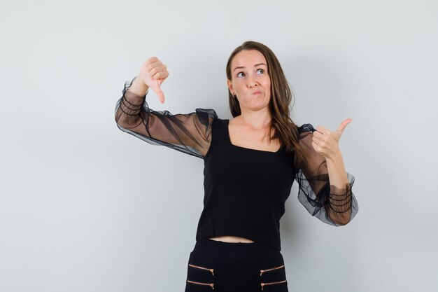 Young woman in black blouse showing thumb up and down and looking puzzled 