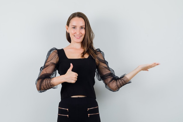 Young woman in black blouse showing thumb up while spreading open palm aside and looking joyful 