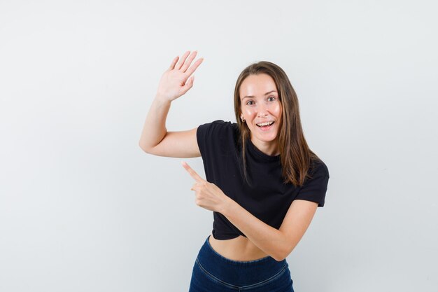 Young woman in black blouse waving hand for greeting while pointing aside and looking joyful