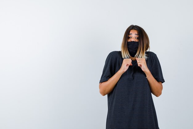 Young woman in black dress, black mask clenching fists and looking scared , front view.