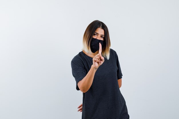 Young woman in black dress, black mask showing hold on a minute gesture, holding hand behind waist and looking happy , front view.