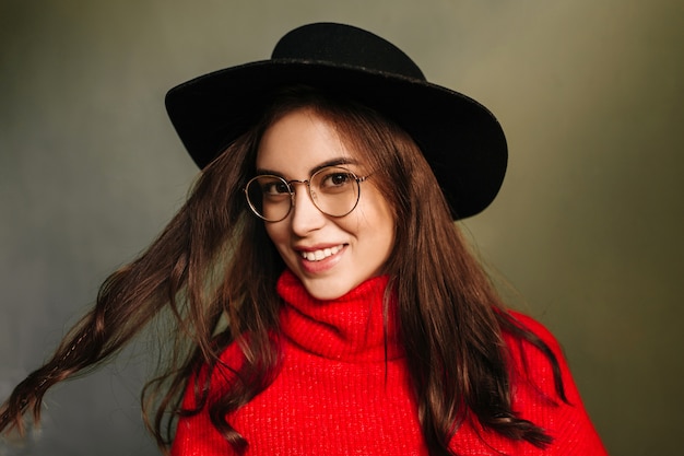 黒い帽子と赤いセーターの若い女性はかわいい笑顔と黒髪で遊ぶ。