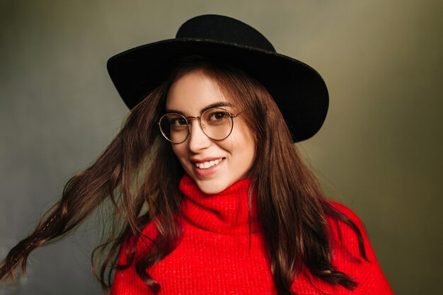 Young woman in black hat and red sweater cute smiles and plays with dark hair.