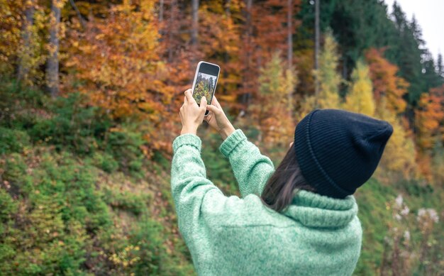 Молодая женщина в черной шляпе фотографирует осенний лес на смартфон