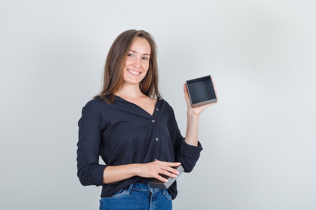 Young woman in black shirt, jeans shorts holding empty watch box and looking cheery