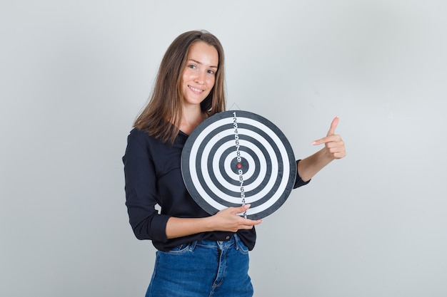 Foto gratuita giovane donna in camicia nera, pantaloncini di jeans che punta il dito al bersaglio e sembra speranzosa