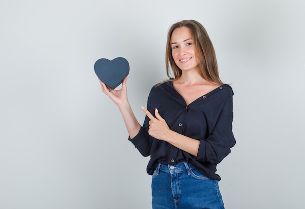 Giovane donna in camicia nera, pantaloncini di jeans che punta il dito alla confezione regalo e sembra gioiosa