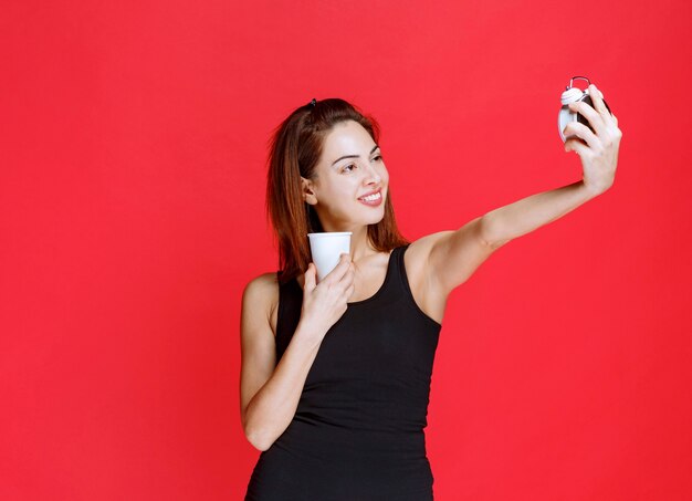 Young woman in black singlet holding an alarm clock and a cup of drink