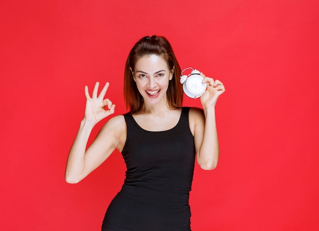 Young woman in black singlet holding an alarm clock and showing positive hand sign