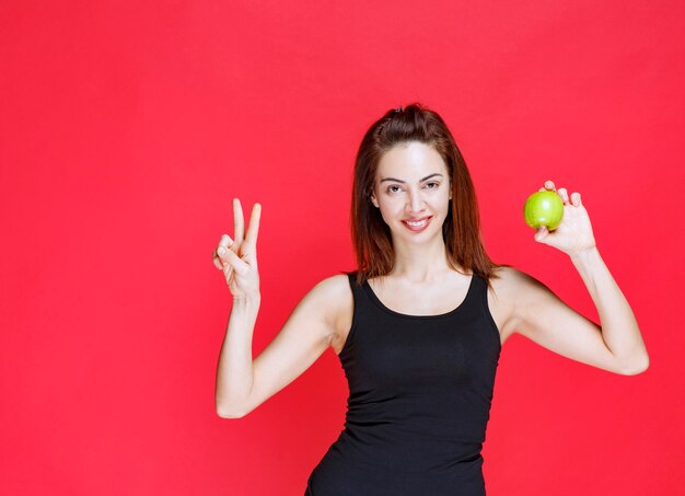 Young woman in black singlet holding green apples and showing peace sign