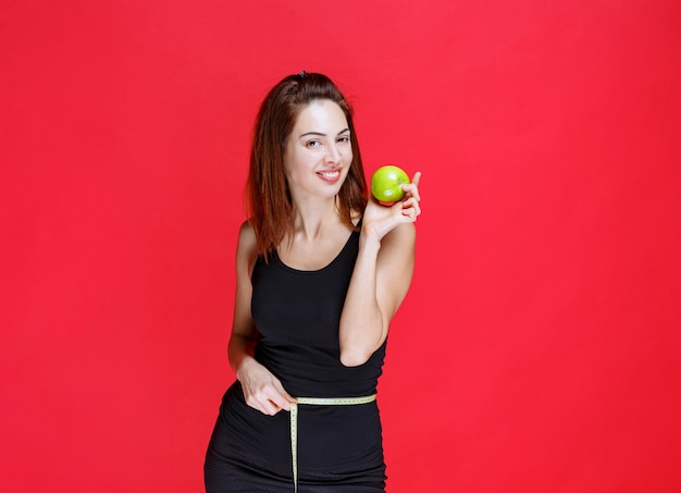 Молодая женщина в черной майке держит зеленые яблоки с измерительной лентой на талии