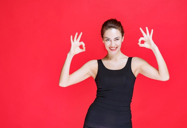 Young woman in black singlet standing on red wall and showing ok sign