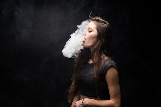 Young woman in black smokes an electronic cigarette on dark wall