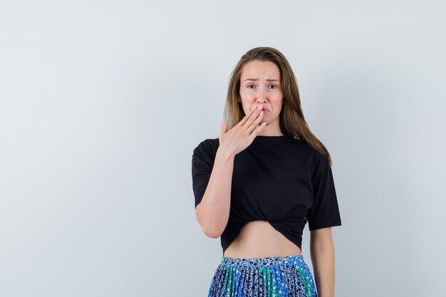 Young woman in black t-shirt and blue skirt covering mouth with hand and looking annoyed