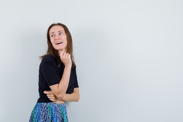 Young woman in black t-shirt and blue skirt looking away while posing at camera and looking happy