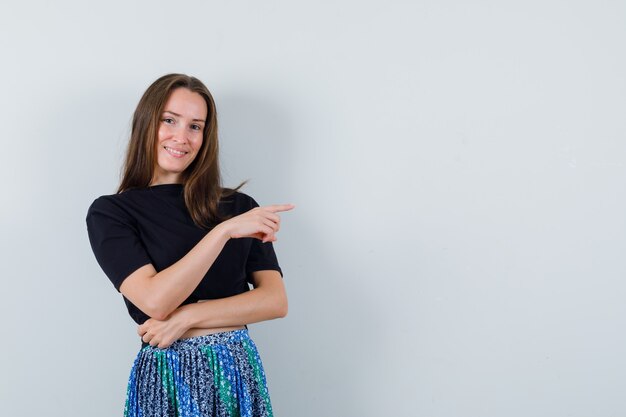 Young woman in black t-shirt and blue skirt pointing right with index finger and looking attractive