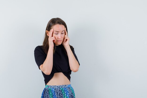 Young woman in black t-shirt and blue skirt rubbing temples and looking tired