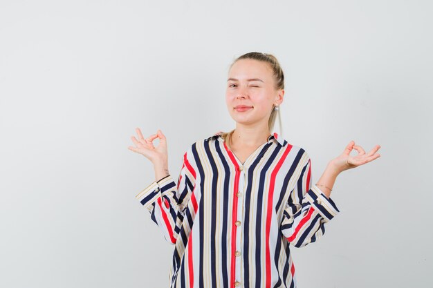 Young woman blinking and showing peace gesture in striped blouse and looking happy