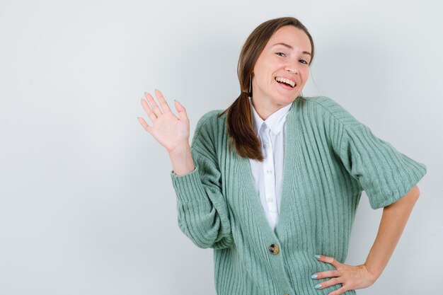 Young woman in blouse, cardigan greeting with open hand and looking jovial , front view.