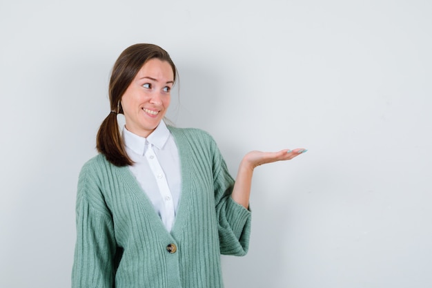 Young woman in blouse, cardigan pretending to hold something, looking aside and looking jolly , front view.