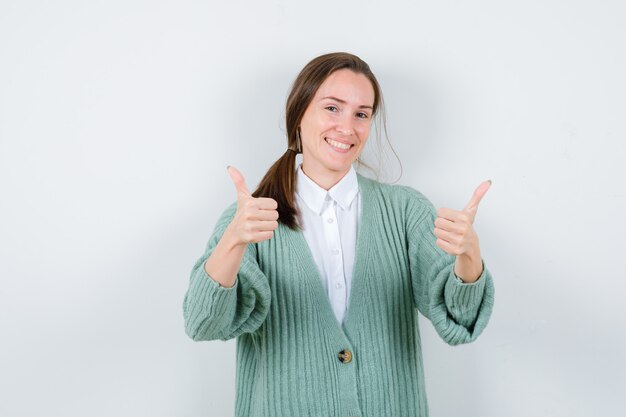 Young woman in blouse, cardigan showing double thumbs up and looking merry , front view.