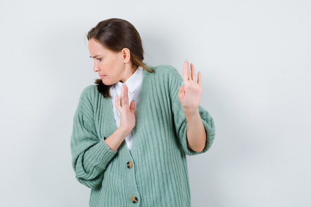 Young woman in blouse, cardigan trying to block herself with hands and looking scared , front view.