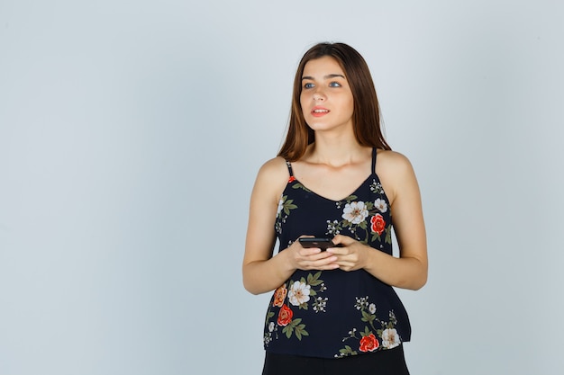 Free photo young woman in blouse holding smartphone while looking away and looking pensive , front view.