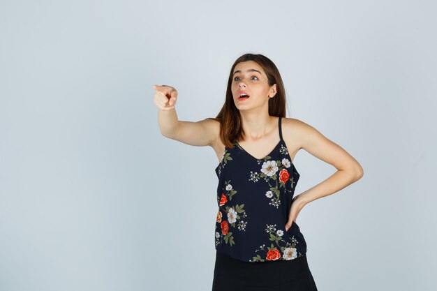 Young woman in blouse, skirt pointing away and looking wondered , front view.