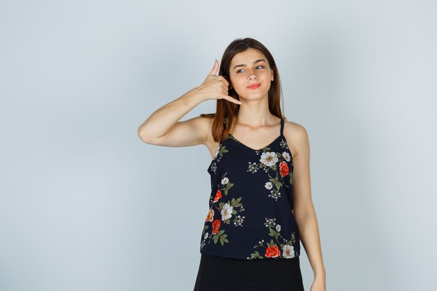 Young woman in blouse, skirt showing phone gesture and looking hopeful , front view.