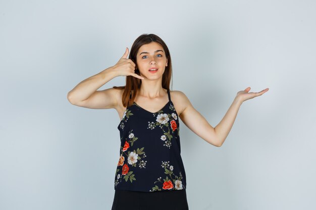 Young woman in blouse, skirt showing phone gesture, spreading palm aside and looking hopeful , front view.