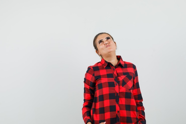 Free Photo young woman blowing cheeks, looking up in checked shirt and looking hopeful. front view.