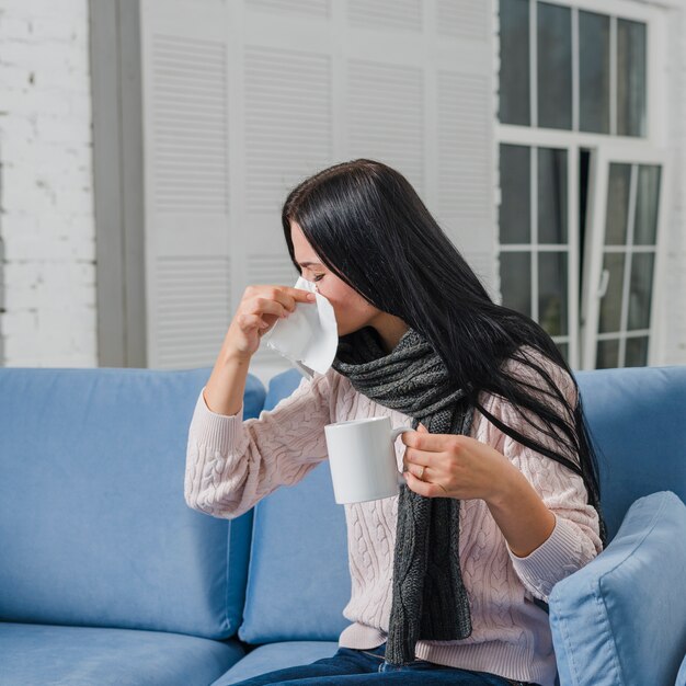 コーヒーのマグカップを手にしてティッシュペーパーで彼女の鼻を吹く若い女性