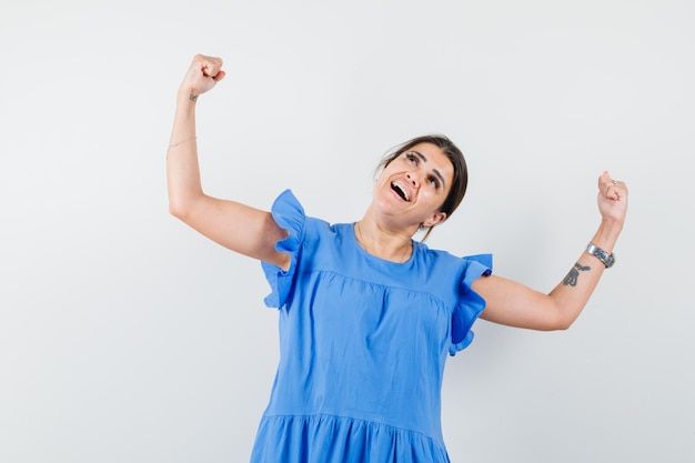 Молодая женщина в синем платье показывает жест победителя и выглядит счастливой