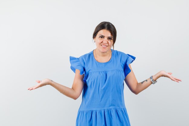 Молодая женщина в синем платье разводит руками в растерянном жесте