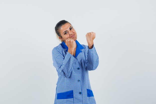 Young Woman In Blue Gingham Pajama Shirt Clenching Fists And Looking Pretty , Front View.