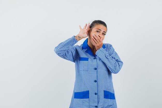 Young woman in blue gingham pajama shirt holding hand near ear as listening to someone and covering mouth with hand and looking surprised , front view.