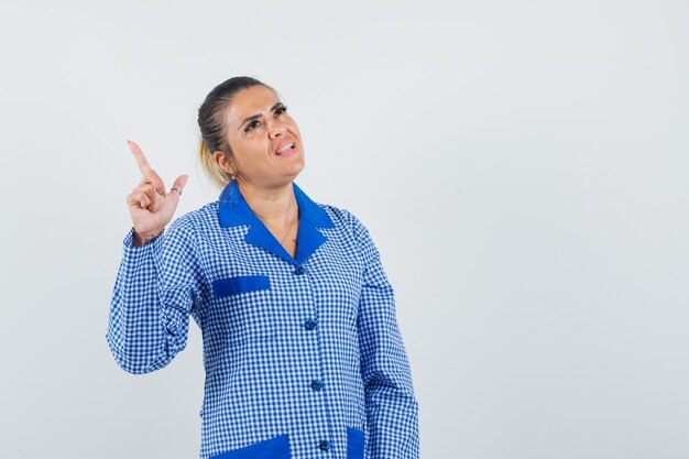 Young woman in blue gingham pajama shirt pointing up and looking pensive , front view.