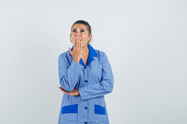 Young woman in blue gingham pajama shirt putting hand on mouth, standing in thinking gesture and looking pensive , front view.