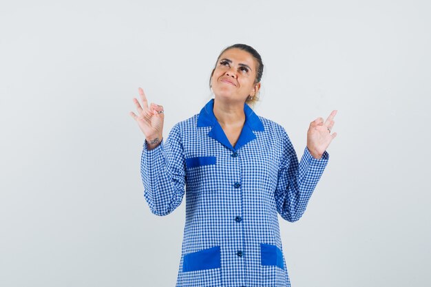Young woman in blue gingham pajama shirt showing ok sign with both hands and looking pretty , front view.