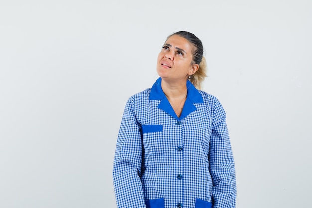 Young woman in blue gingham pajama shirt standing straight while looking above and looking pensive , front view.