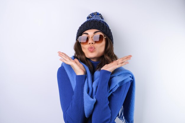 파란색 골프 터틀넥 모자와 흰색 배경에 스카프 선글라스를 쓴 젊은 여성이 기분이 좋습니다.